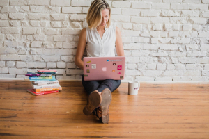 Girl sat on wooden floor leaning against white-painted brick wall. Working on pink laptop with a pile of books on her right and a coffee cup to her left.